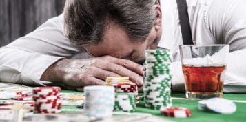 Depressed man leaning his head at a casino table, with chips and alcohol in front him.